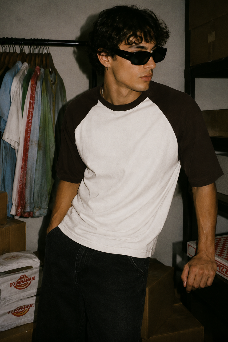 A man wearing a brown and white raglan t-shirt and black sunglasses, standing in a storage room with cardboard boxes and a clothing rack in the background.