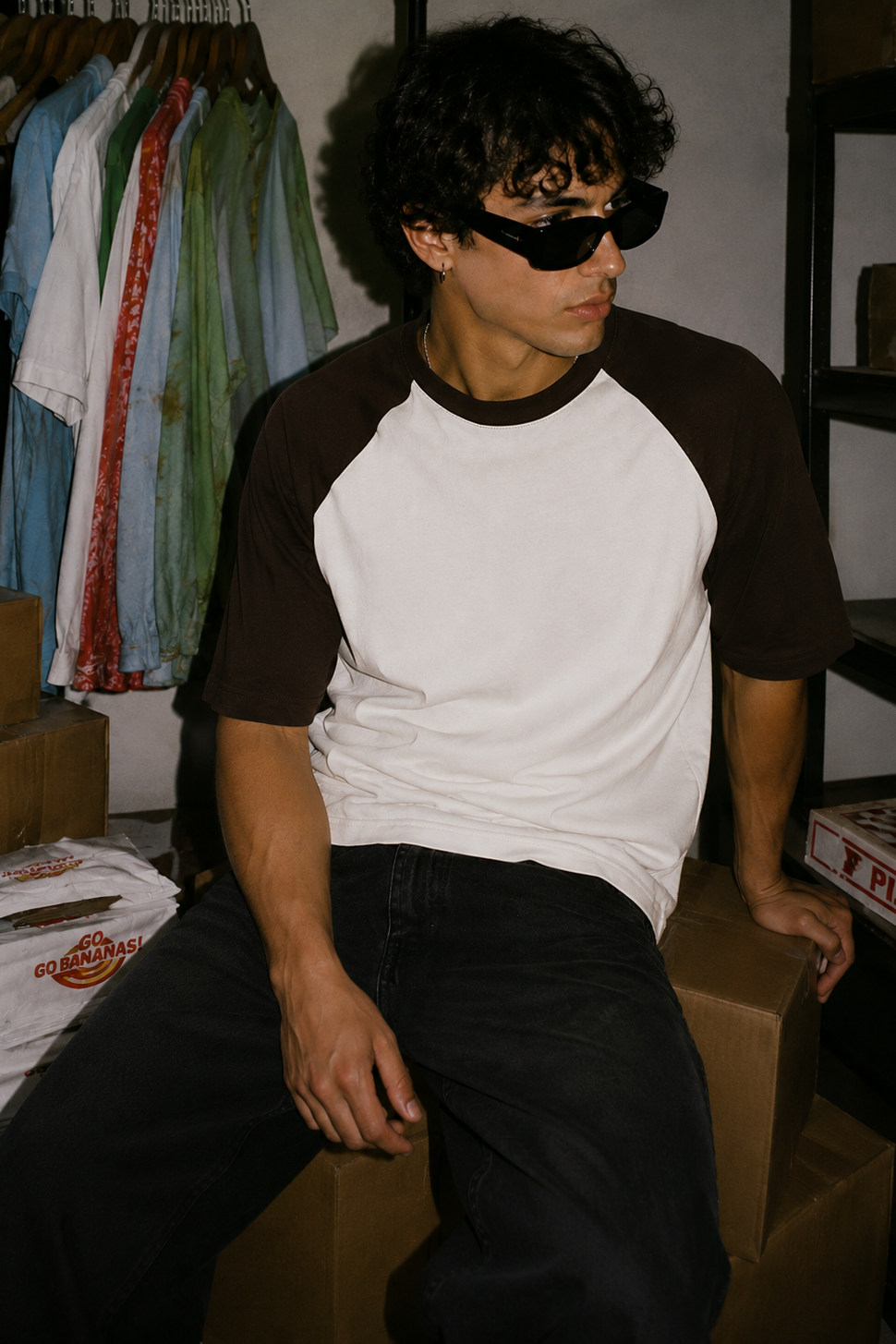 A man wearing a white and dark brown raglan t-shirt, black sunglasses, and black jeans sits on cardboard boxes in a storage room with clothes hanging in the background.