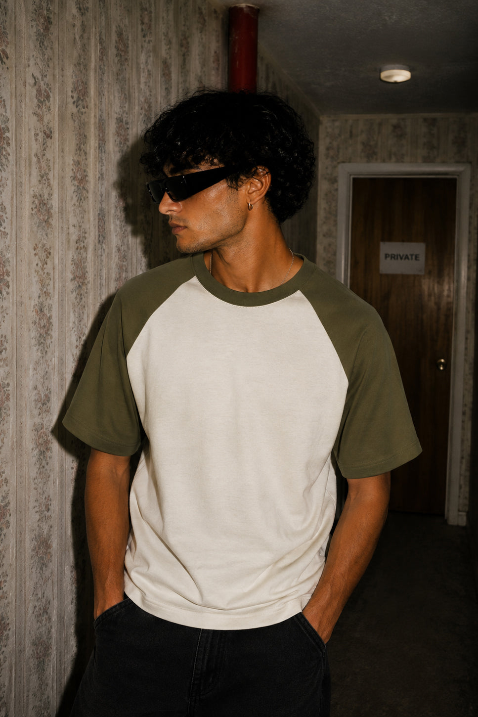 A man wearing a short-sleeve raglan t-shirt with an off-white body and olive green sleeves, styled with black sunglasses and dark pants in a dimly lit hallway.
