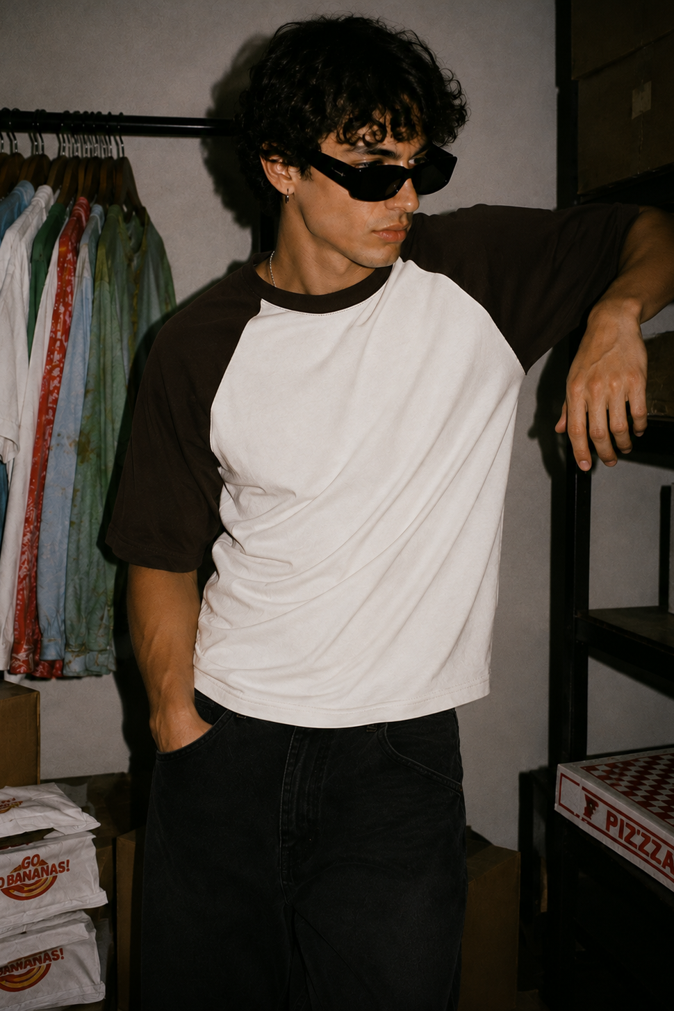 A person wearing a white and dark brown raglan t-shirt, black sunglasses, and black jeans, leaning against a shelf in a storage room with clothes and pizza boxes in the background.