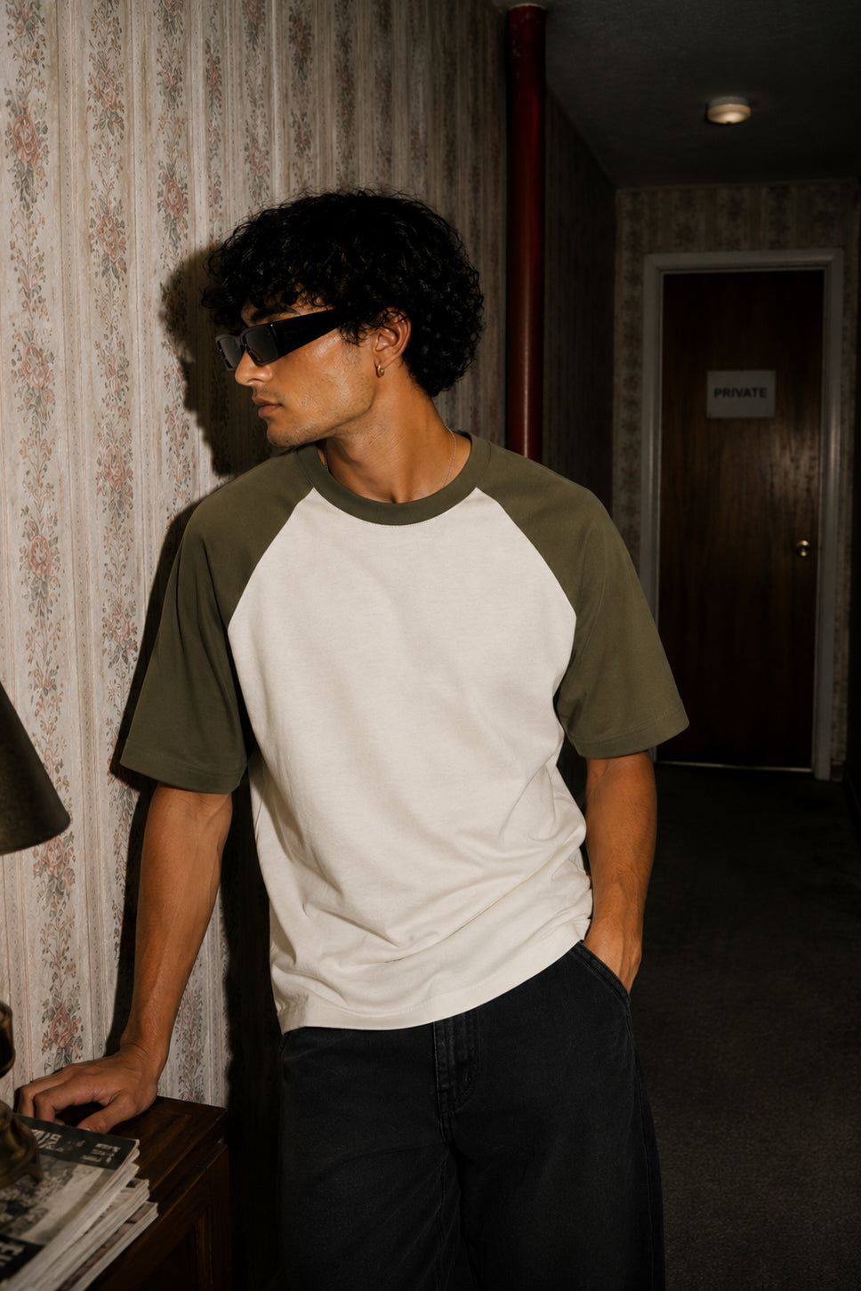 A man wearing a white and olive green raglan t-shirt, black sunglasses, and dark pants, standing in a dimly lit hallway with patterned wallpaper.