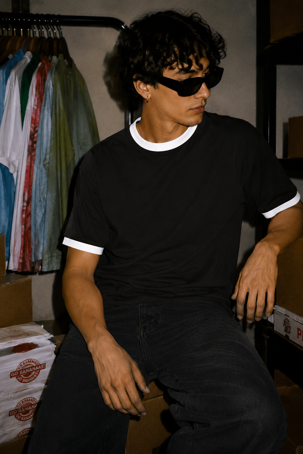 A man wearing a black ringer t-shirt with white trim on the crew neck and sleeve cuffs, paired with black jeans and black sunglasses, sitting in a storage room with cardboard boxes and a clothing rack in the background.