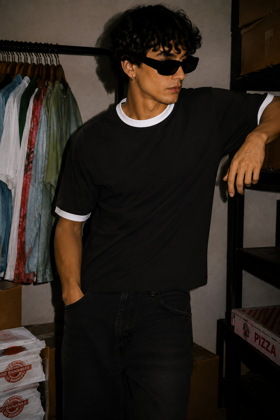 A man wearing a black ringer t-shirt with white ribbed collar and sleeve cuffs, black jeans, and black sunglasses, leaning against a shelf in a storage room setting.