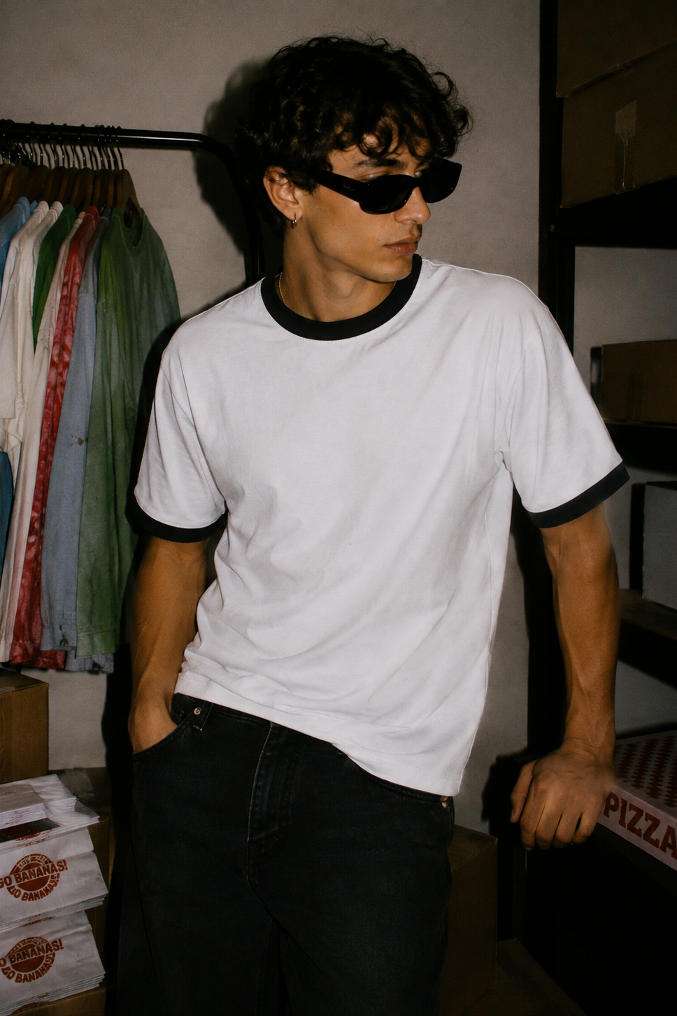 A man wearing a white ringer t-shirt with black trim and black sunglasses, standing in a dimly lit room with clothing racks and cardboard boxes in the background.