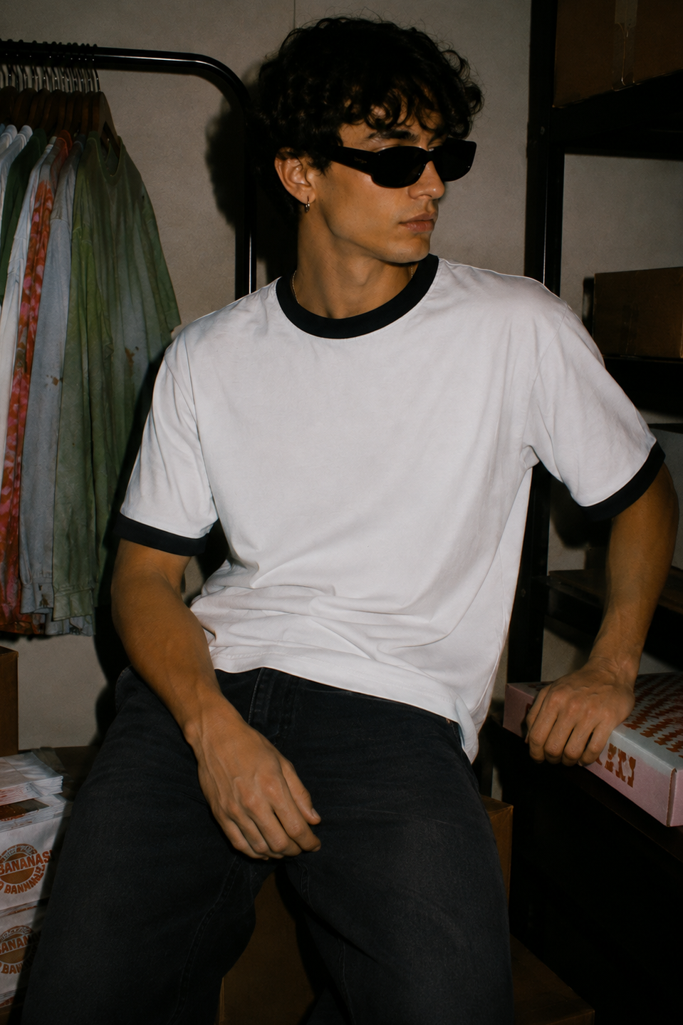 A man wearing a white ringer t-shirt with black trim and black sunglasses, sitting in a storage room with clothing racks and boxes in the background.