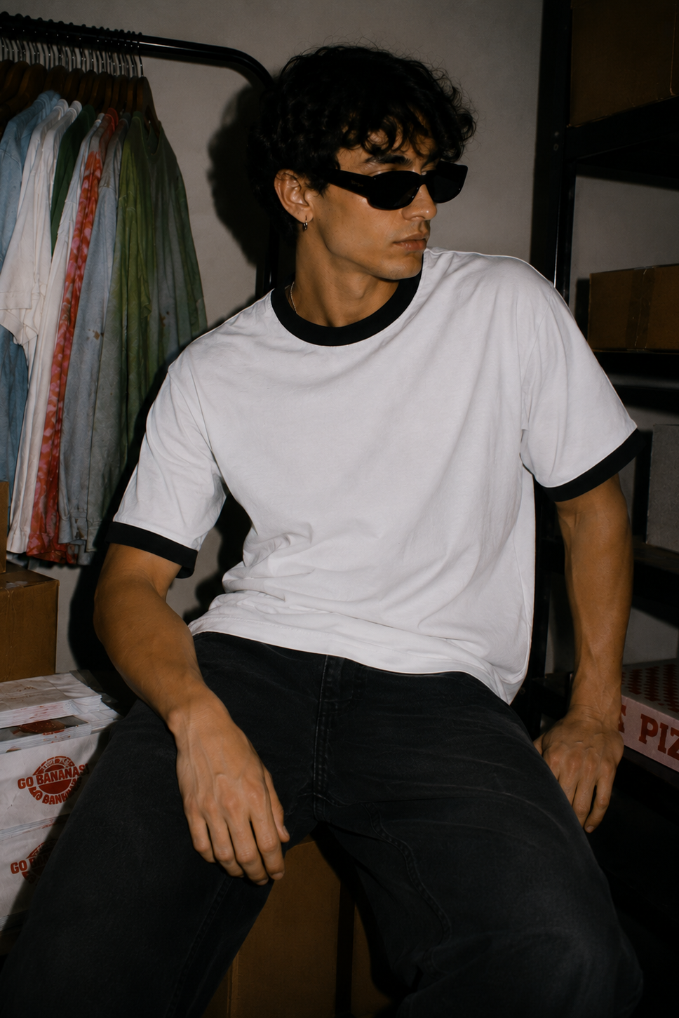 A man wearing a white ringer t-shirt with black trim and black sunglasses, sitting in a storage room with cardboard boxes and a clothing rack in the background.