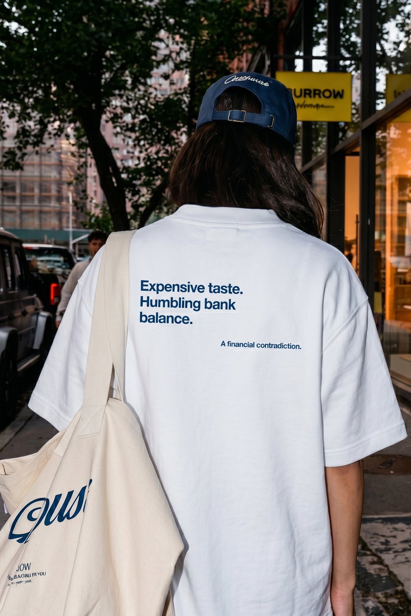 Person wearing a white t-shirt with text and carrying a beige tote bag on a city street.