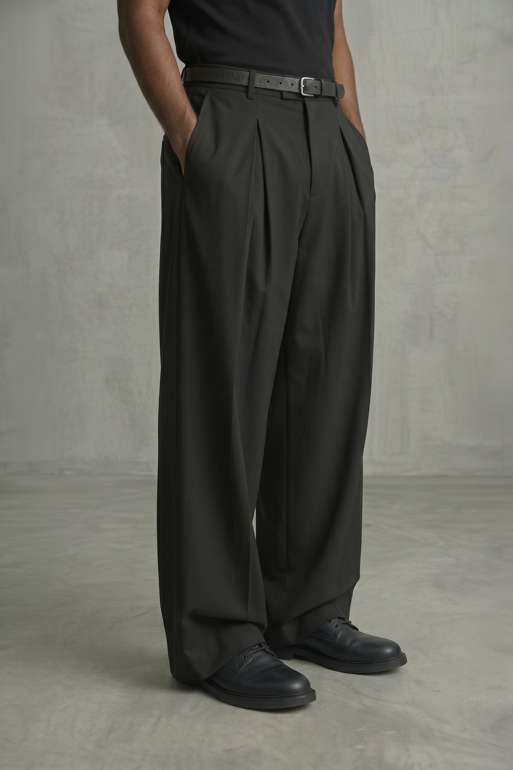 Person wearing dark gray pleated pants and black shoes against a plain background