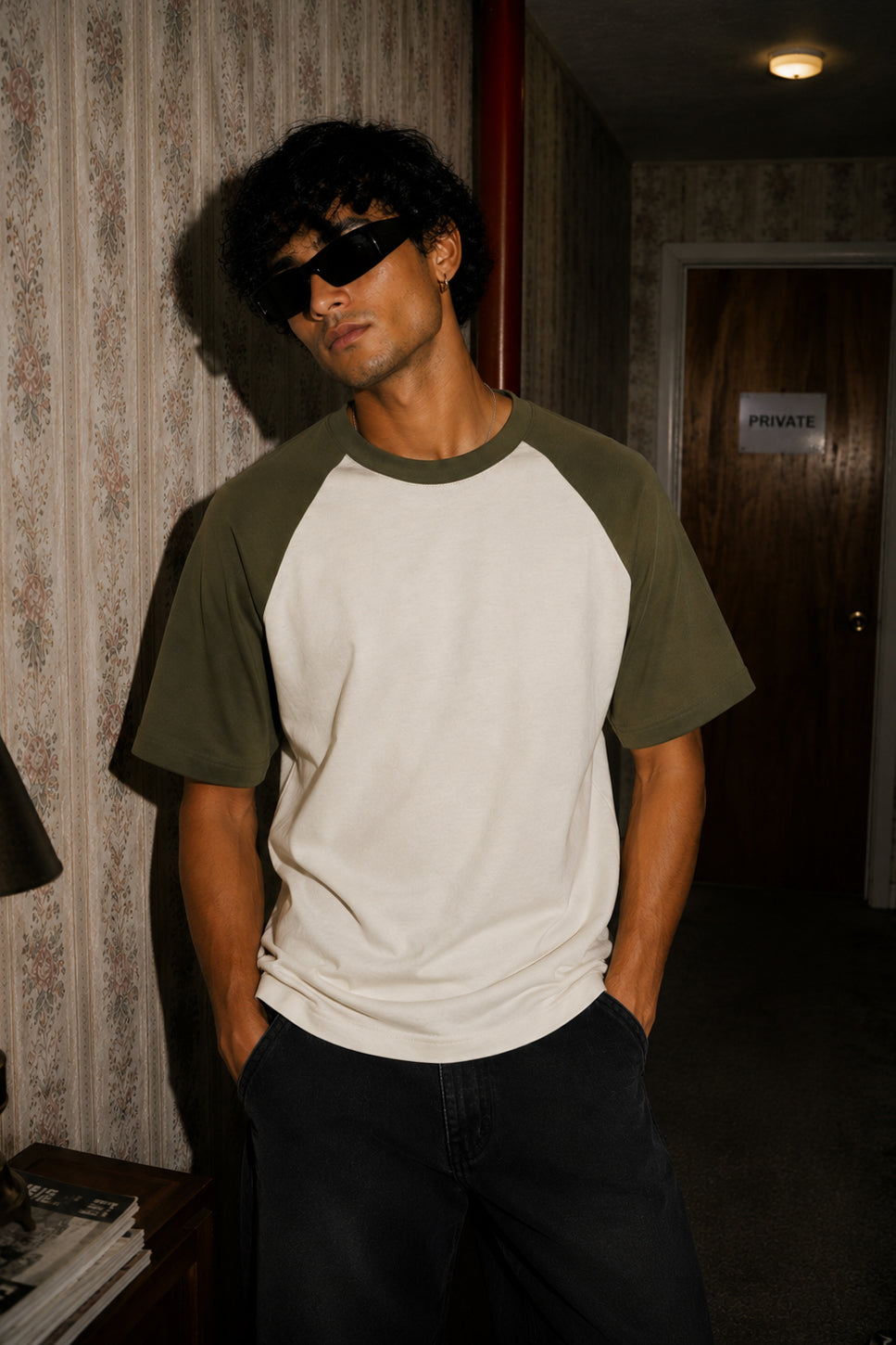 A man wearing a two-tone raglan t-shirt with an off-white body and olive green sleeves, styled with dark sunglasses and dark pants in a dimly lit hallway.