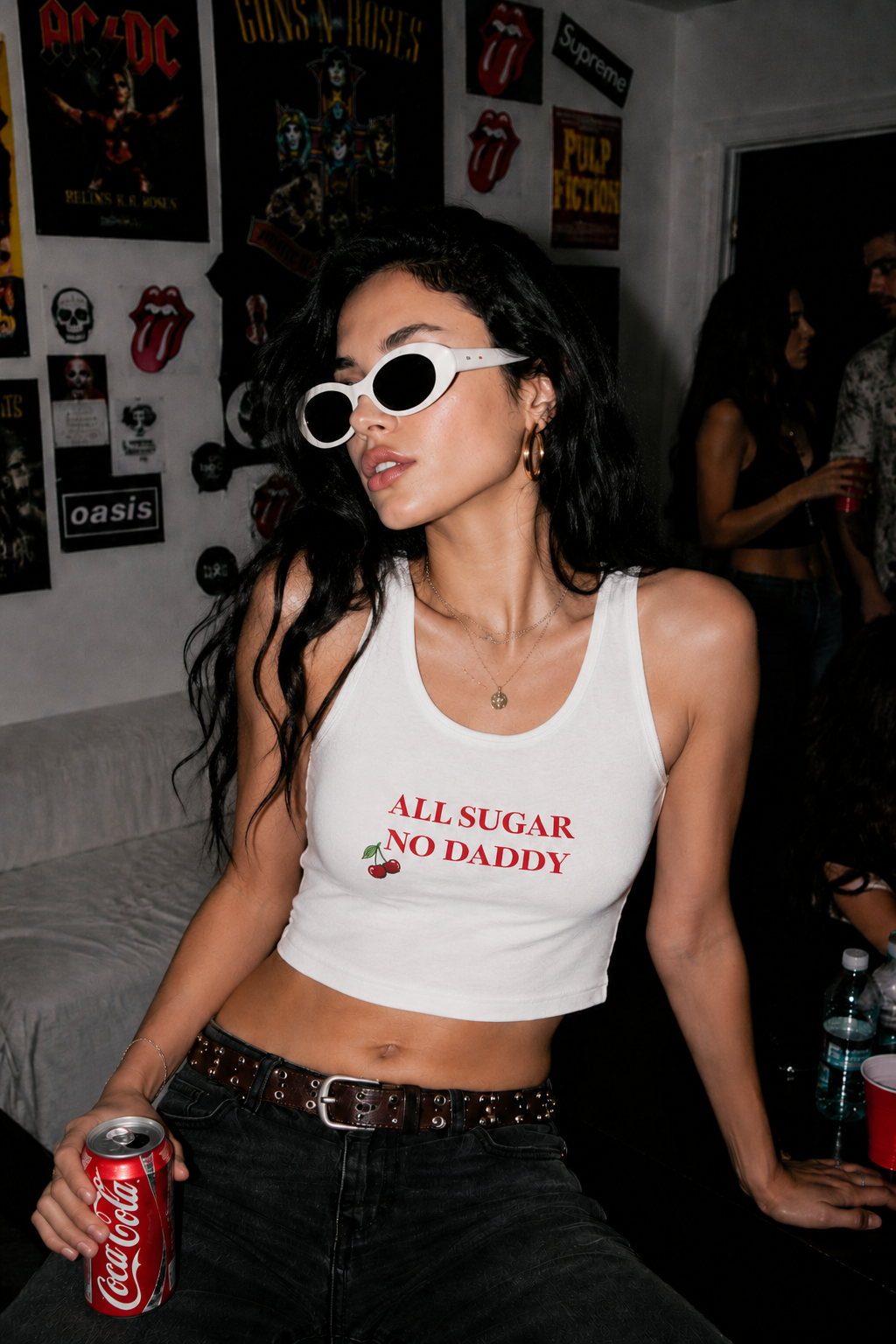 A woman wearing a white cropped tank top with red text reading 'ALL SUGAR NO DADDY' and a small cherry graphic, paired with dark jeans and white-framed sunglasses, holding a Coca-Cola can in a dimly lit room with music posters on the wall.