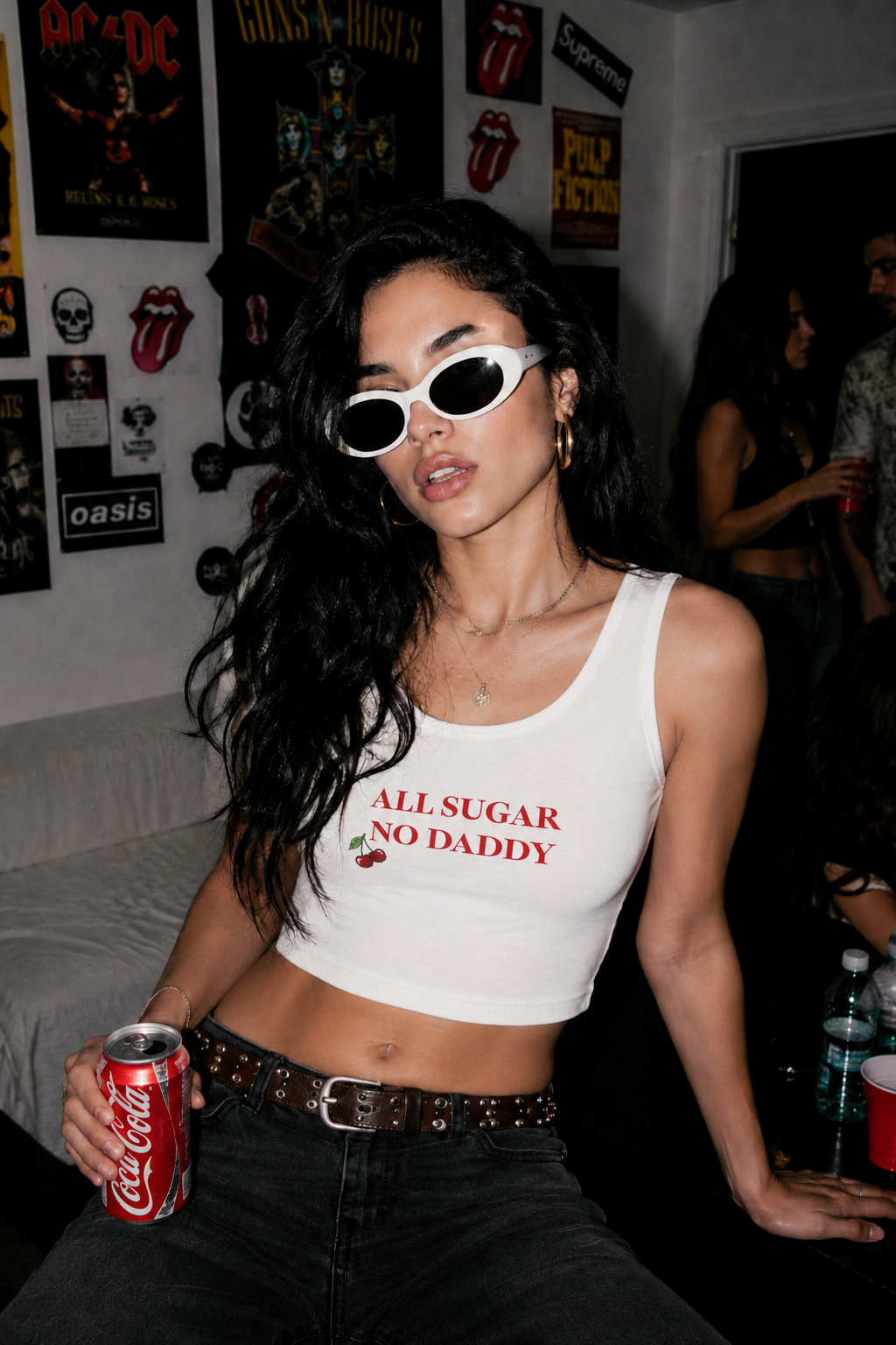 A woman wearing white cat-eye sunglasses and a white crop top with red text reading 'ALL SUGAR NO DADDY' sits in a dimly lit room with posters on the wall. She is holding a red Coca-Cola can.