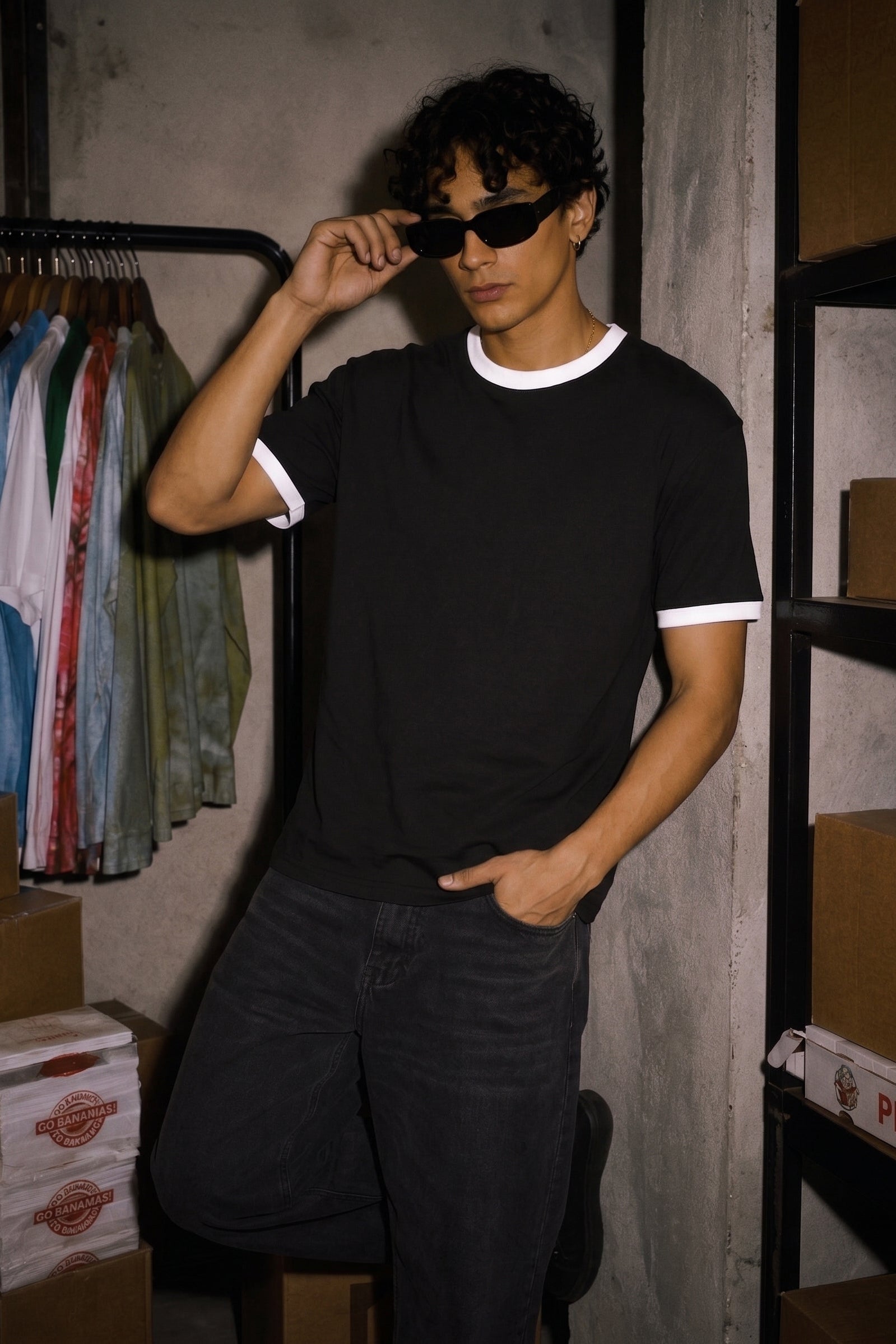 A man wearing a black ringer t-shirt with white trim and dark sunglasses, posing in a dimly lit warehouse or storage room with cardboard boxes and clothing racks in the background.