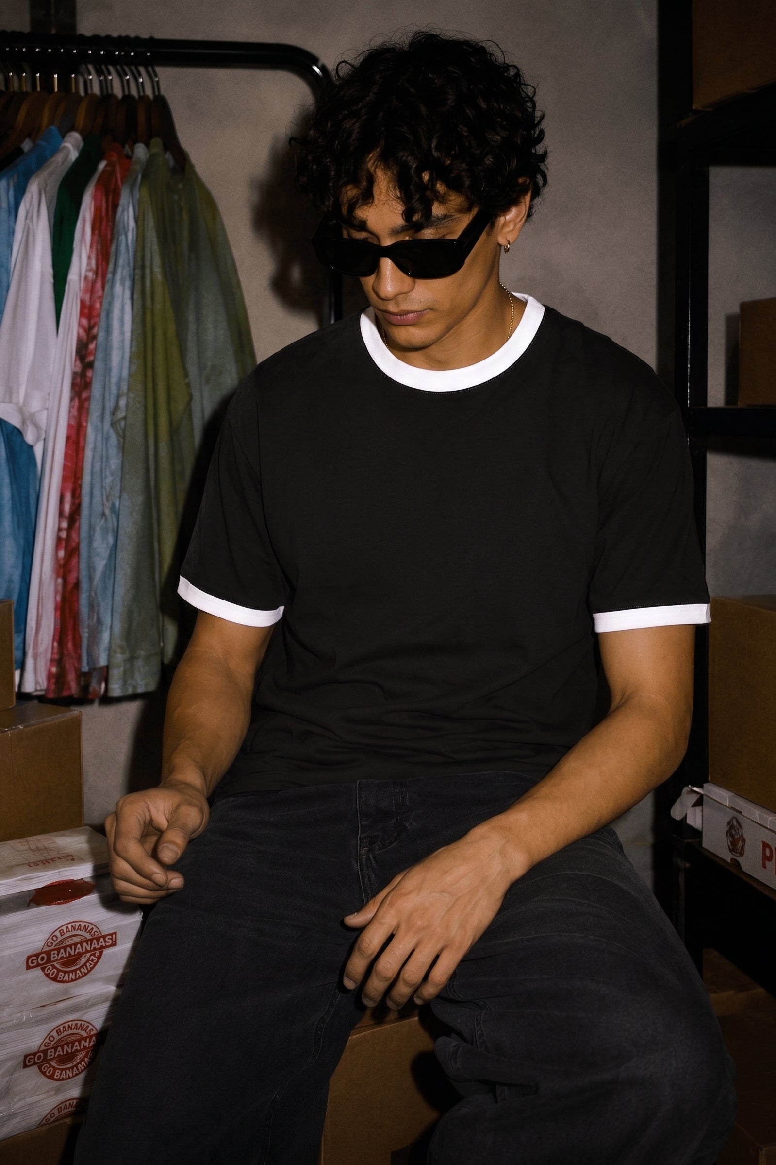 A man wearing a black ringer t-shirt with white trim and dark sunglasses, sitting in a storage room with cardboard boxes and a clothing rack in the background.