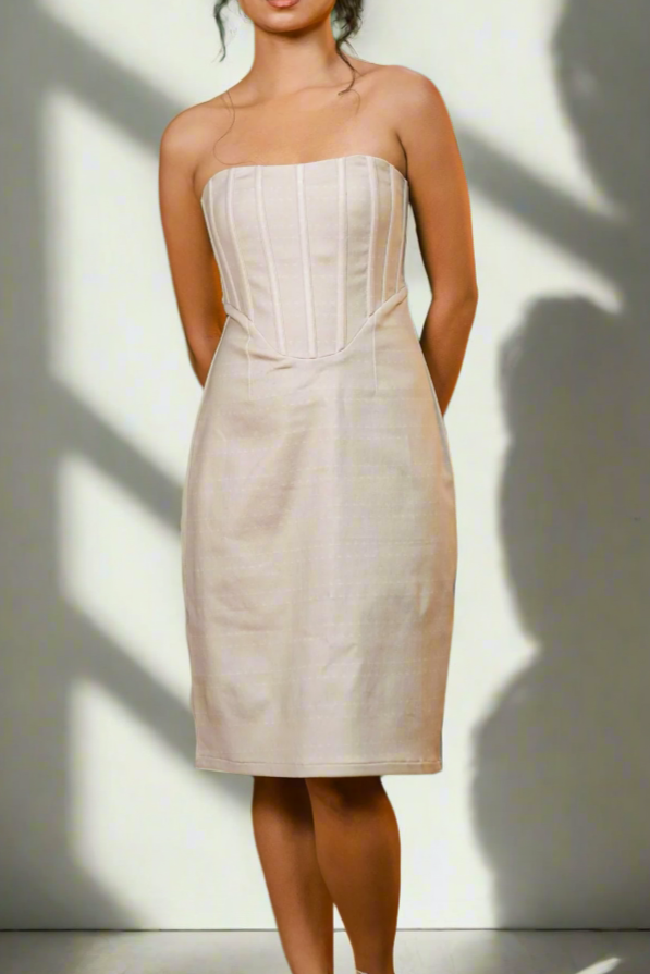 Woman wearing a strapless beige dress standing against a light gray wall with a soft shadow.