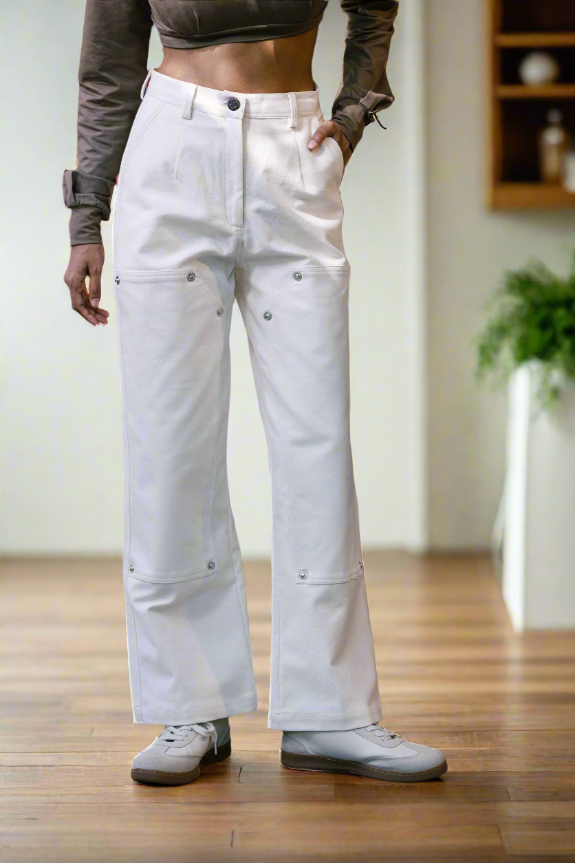 A person standing on a tiled floor wearing white denim pants with a straight silhouette and exaggerated show pockets, paired with gray sneakers.