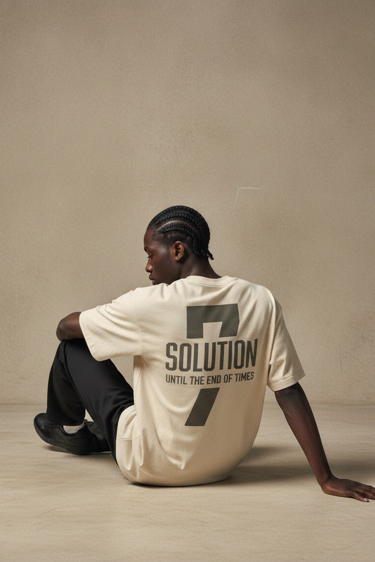 Person wearing a beige t-shirt with text sitting on the floor against a plain wall.