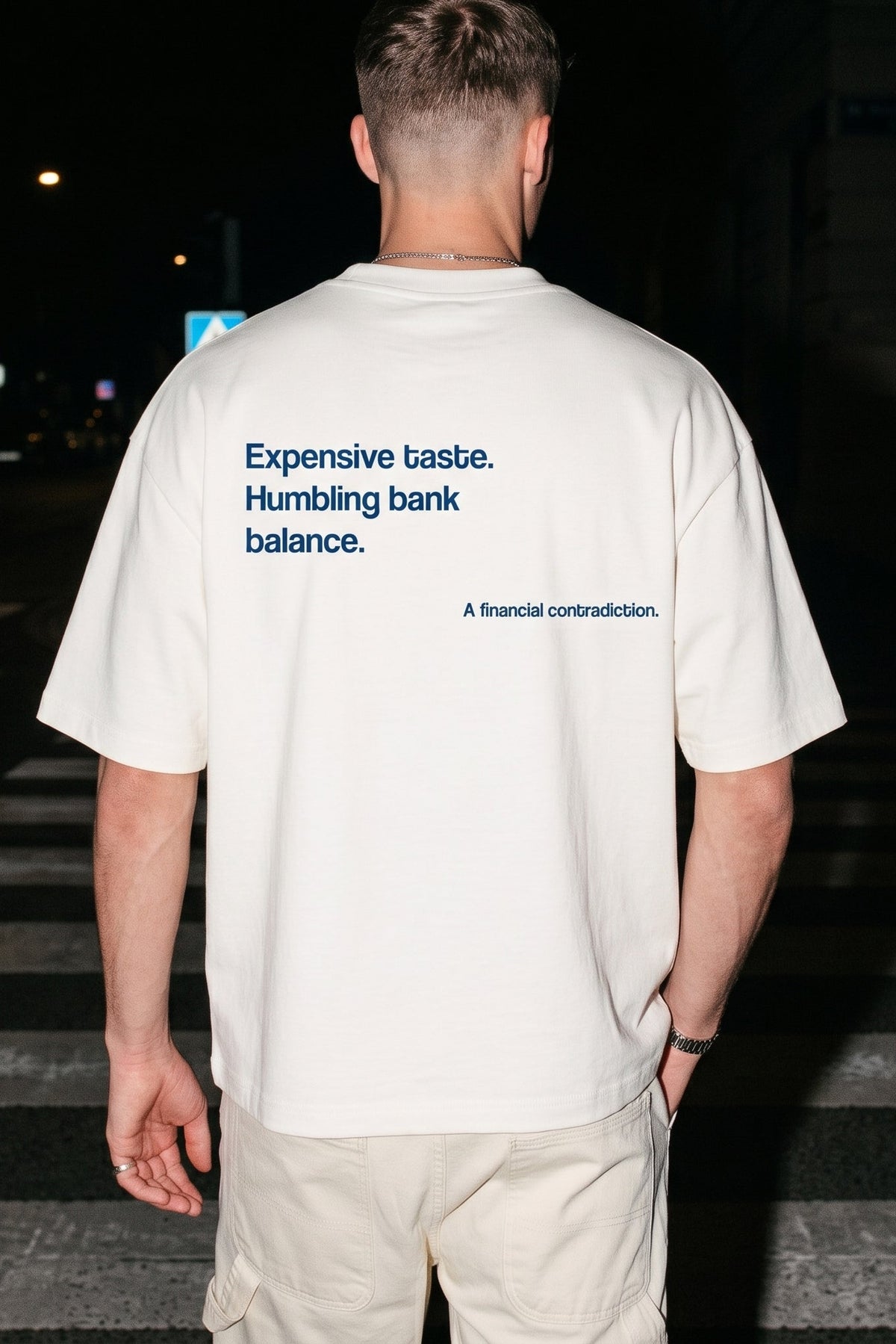 A man viewed from behind wearing an off-white t-shirt with blue text on the back that reads 'Expensive taste. Humbling bank balance.
