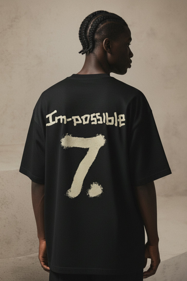 Person wearing a black t-shirt with 'I'm possible 7' text on the back against a beige wall.