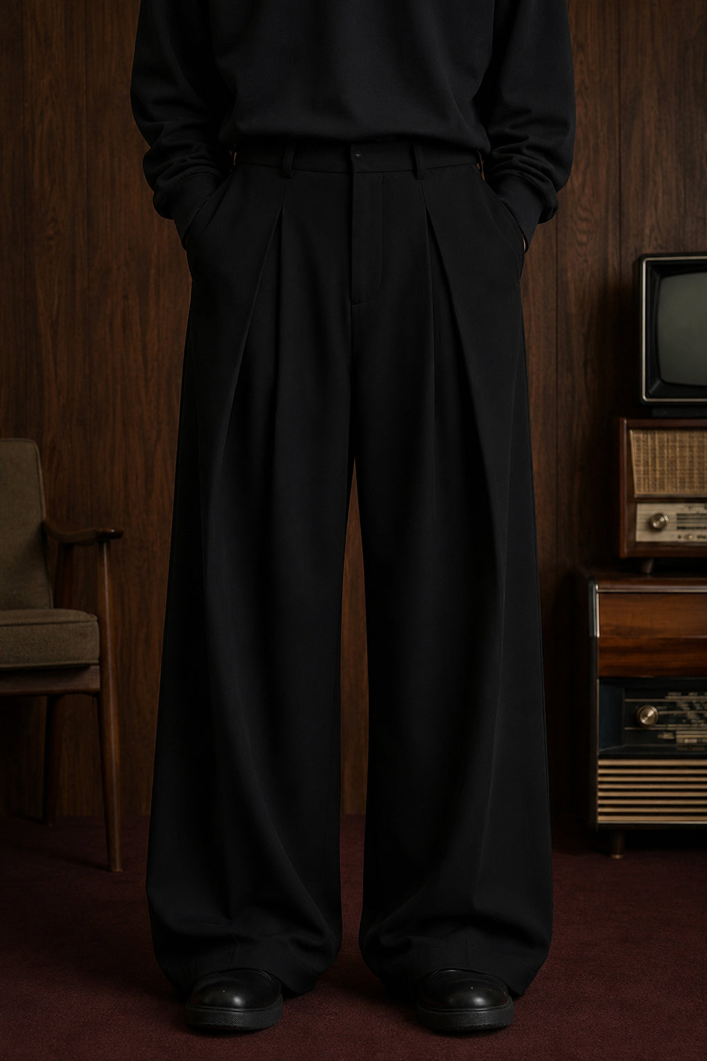 A person wearing black wide-leg pleated trousers with a black long-sleeve top, standing in a vintage room with wood paneling, a small television, and a vintage radio.