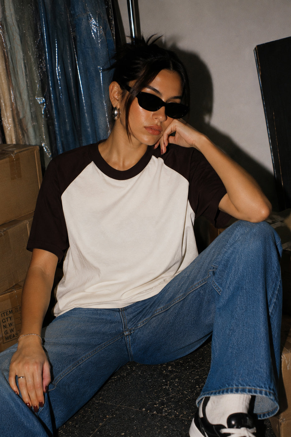 A woman wearing a black and off-white raglan t-shirt, blue jeans, and black sunglasses sits in a dimly lit warehouse-like setting.
