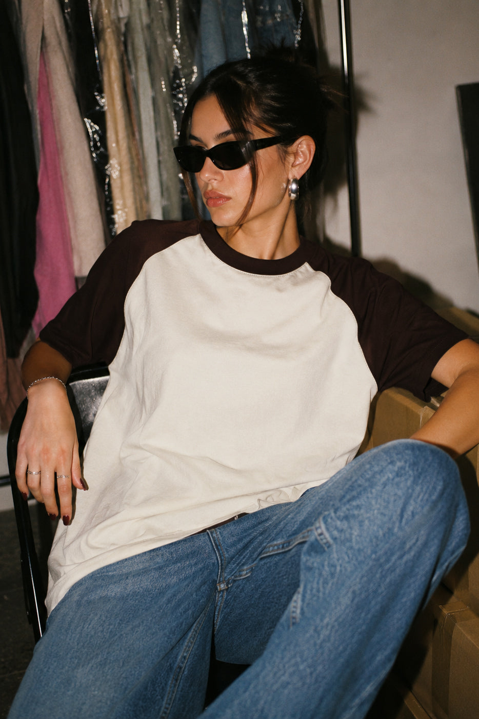 A woman wearing a brown and off-white raglan t-shirt, blue jeans, and black sunglasses sits in a chair in a room with clothing racks in the background.
