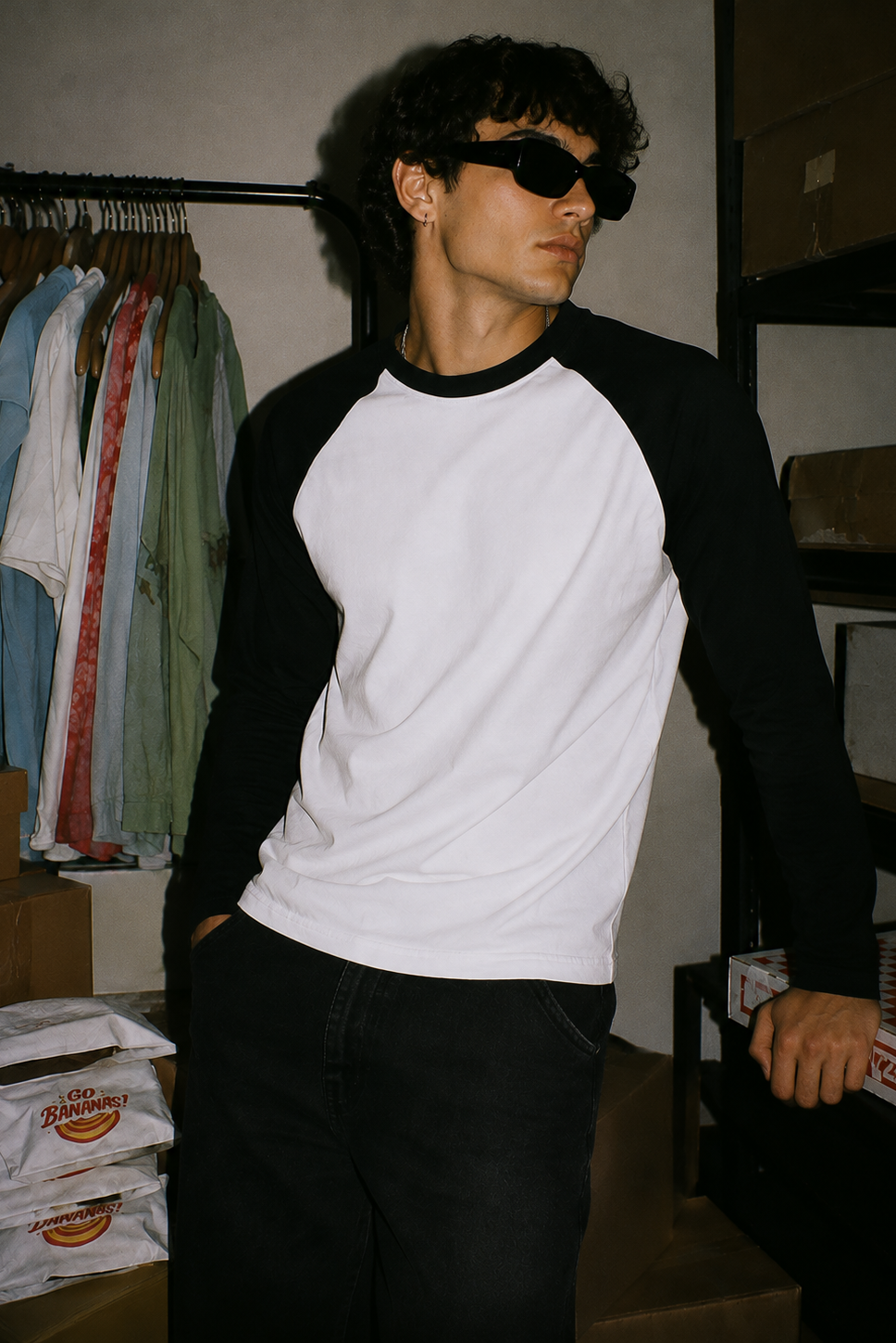 A man wearing a black and white raglan baseball t-shirt, black sunglasses, and black jeans, standing in a storage room with cardboard boxes and a clothing rack in the background.