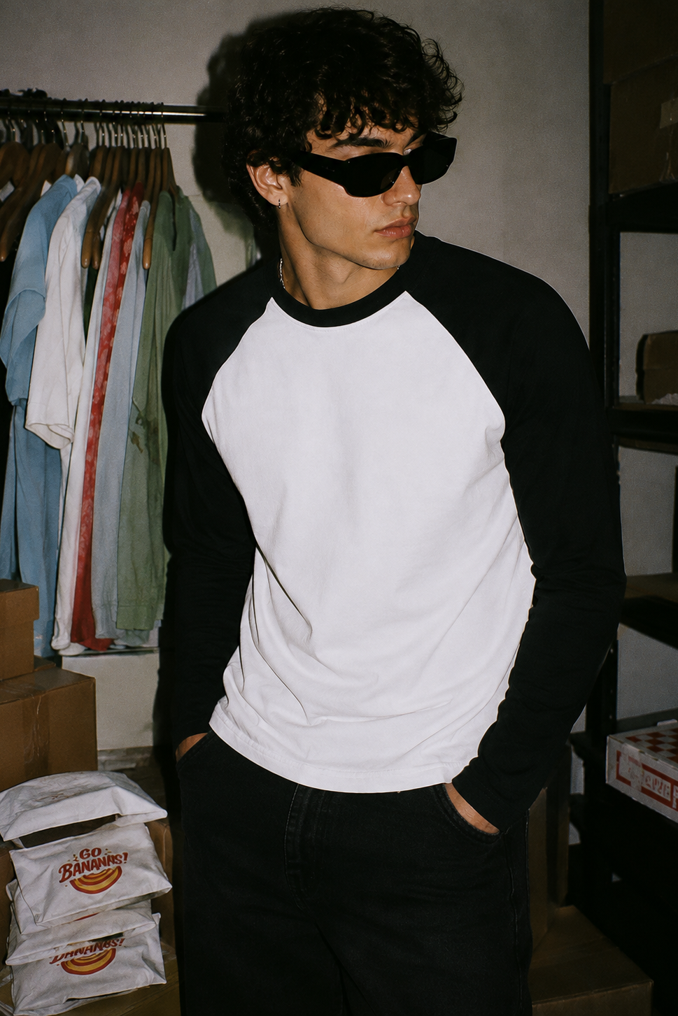 A man wearing a black and white raglan baseball t-shirt and black sunglasses, standing in a storage room with clothes on a rack and cardboard boxes in the background.