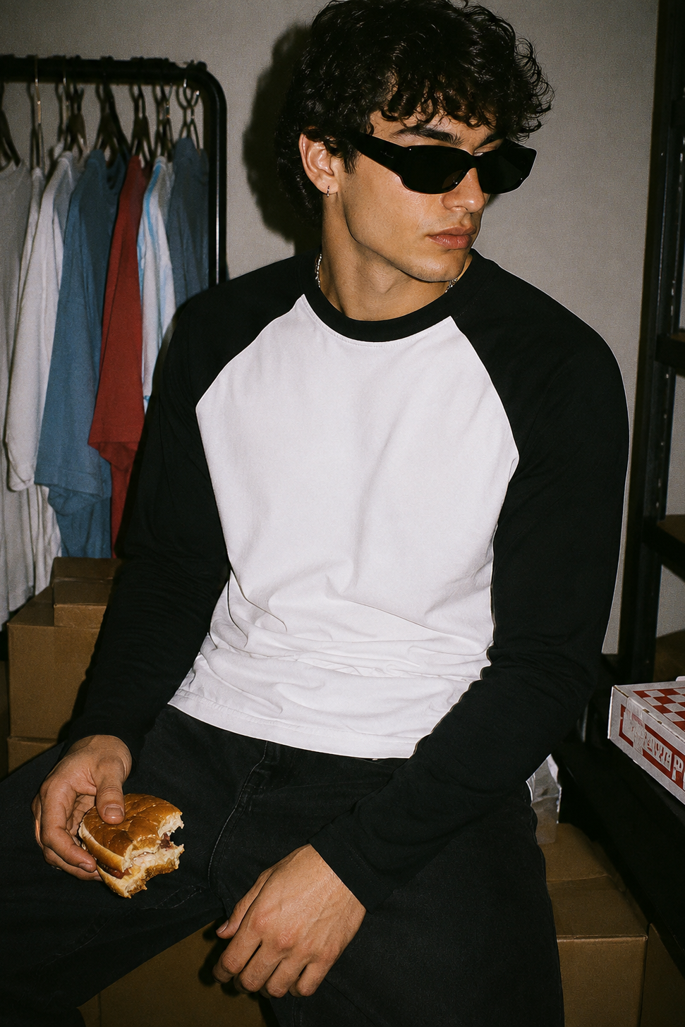 A man wearing a black and white raglan baseball t-shirt and black sunglasses, holding a sandwich while sitting in a room with cardboard boxes and a clothing rack in the background.